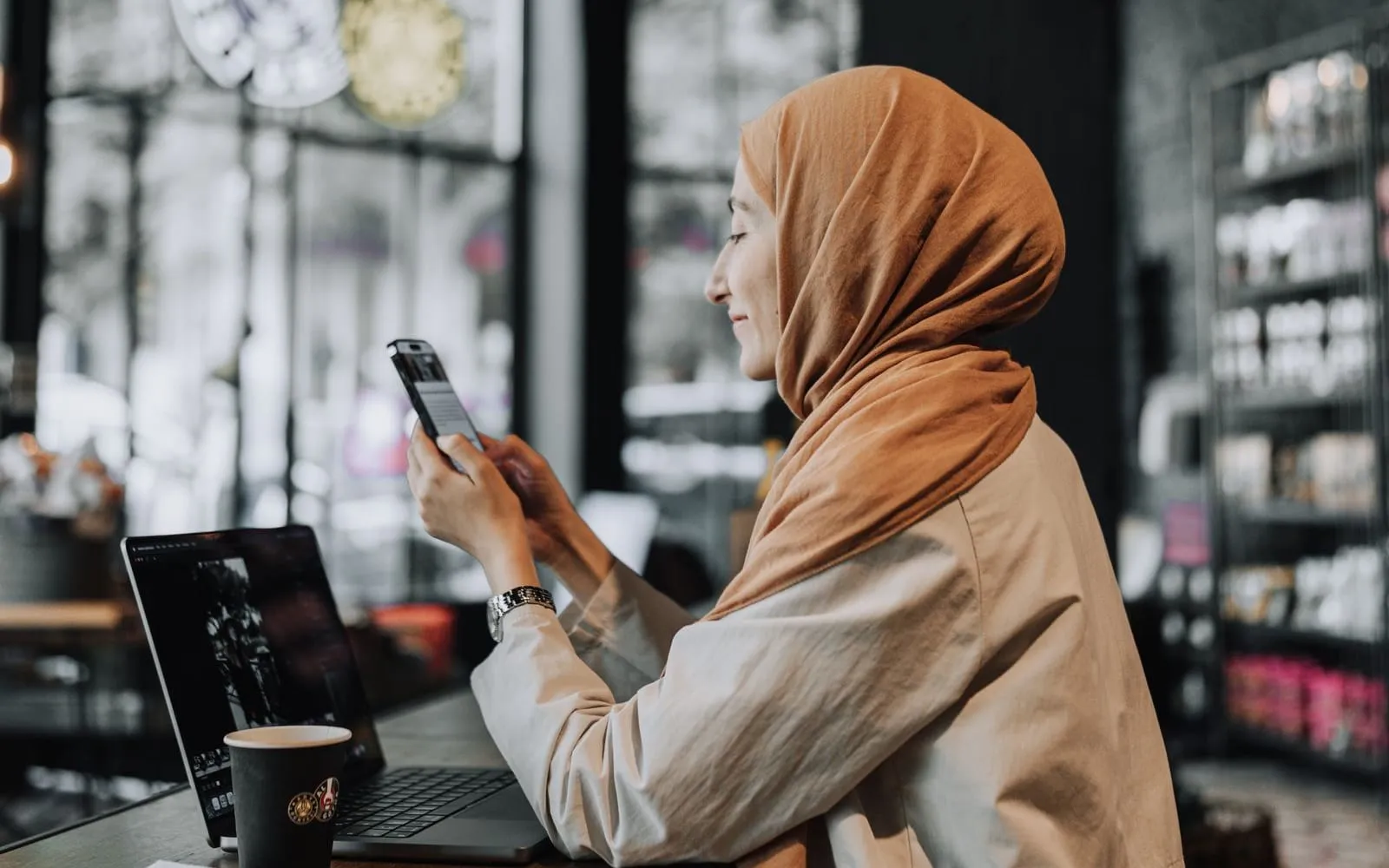 Mulher sentada num café usando o celular, com o notebook na mesa