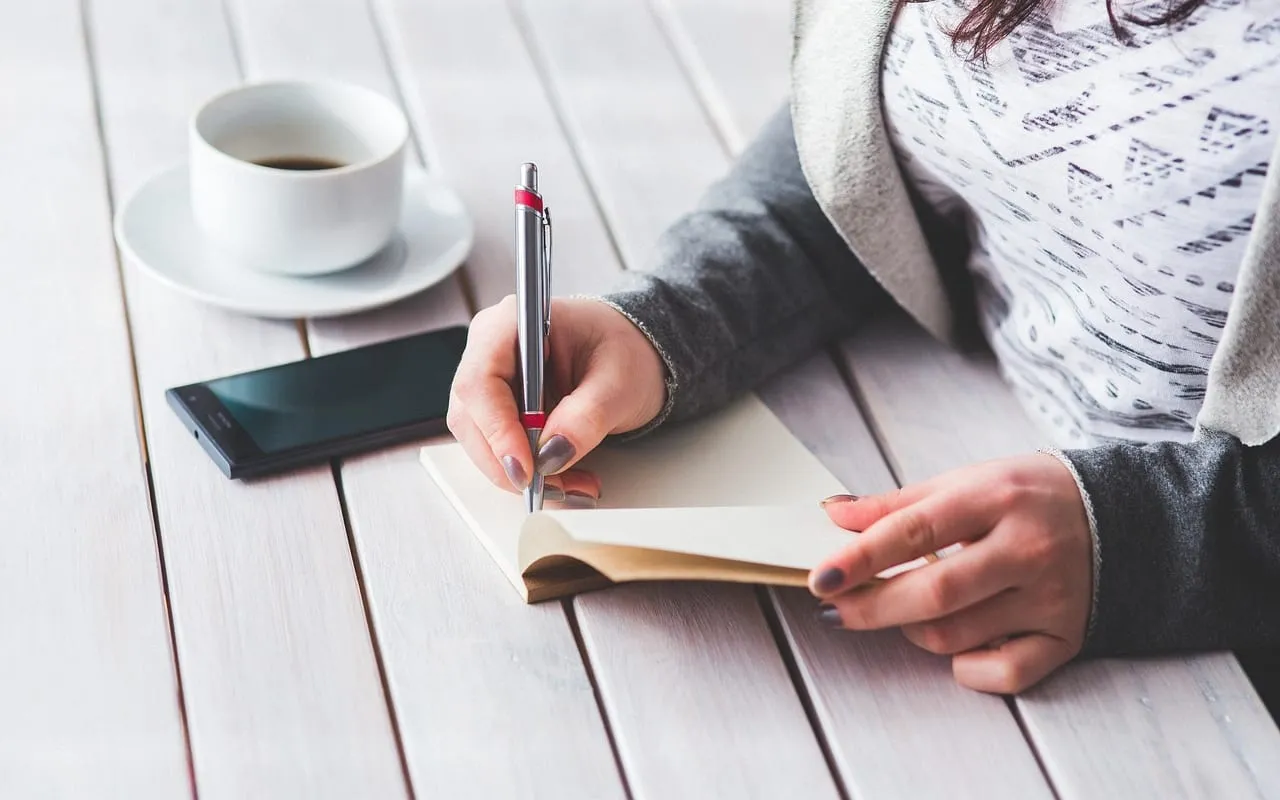 Mulher anotando num caderno, com uma xícara de café e o celular sobre a mesa