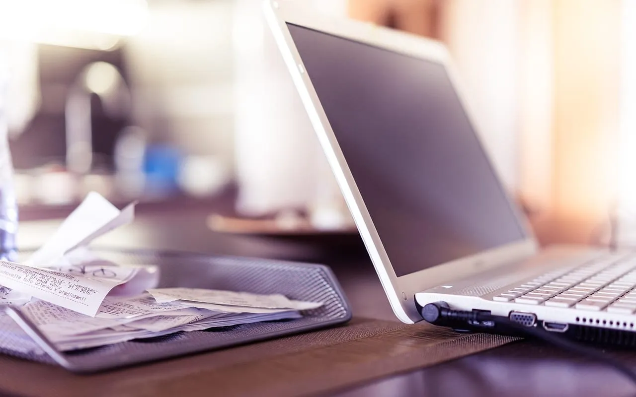 Notebook aberto na mesa, ao lado de uma bandeja com comprovantes de compra