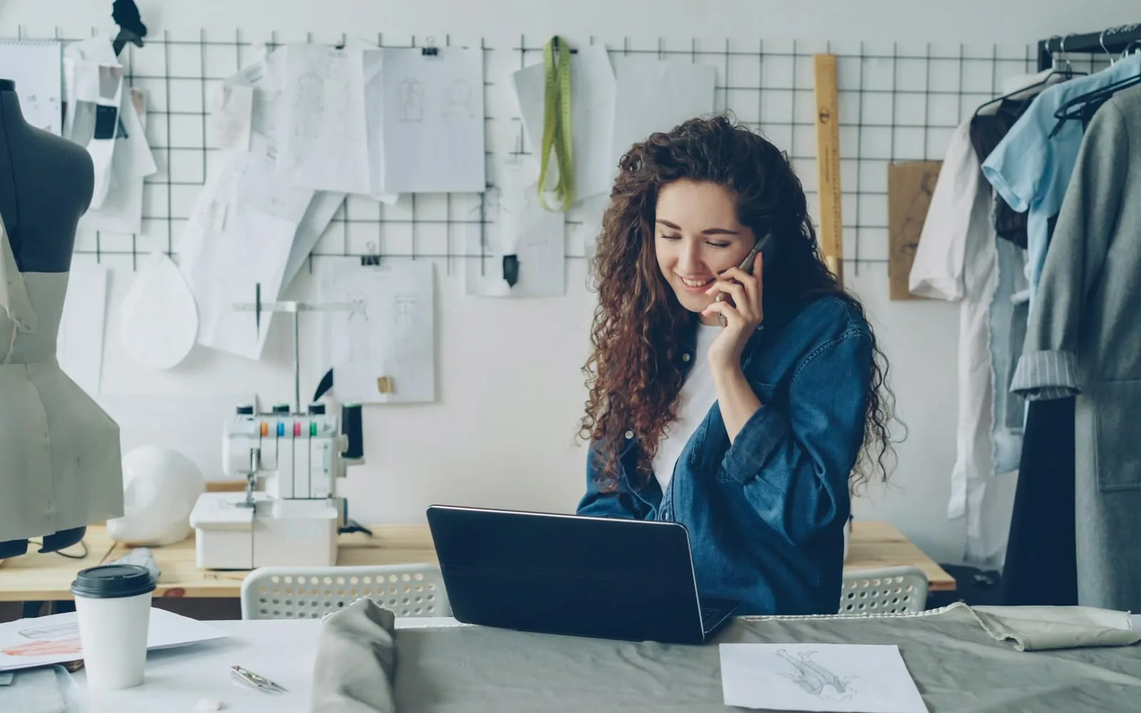 Empreendedora no ateliê falando ao telefone, com o notebook na mesa