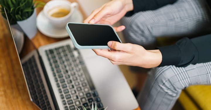 Pessoa consultando aplicativo financeiro no celular ao lado de notebook em ambiente de trabalho